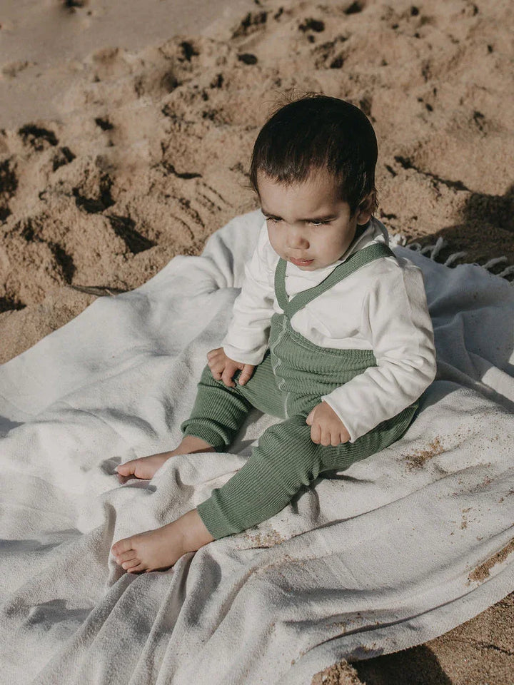 Baby i grønne bukser og hvit genser på sandstrand, myke barneklær for små barn