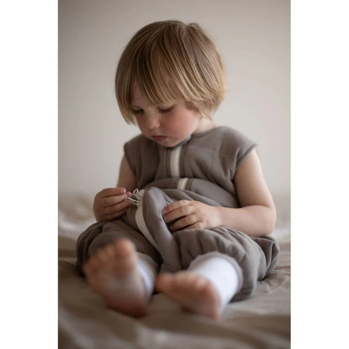 Liten gutt i myk, grå barneklær med hvite detaljer, sitter på seng