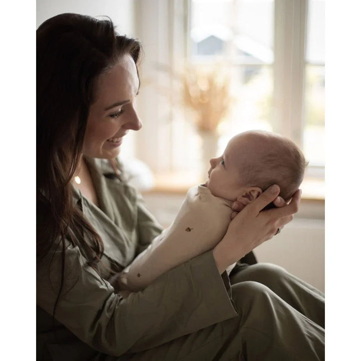 Mor holder nyfødt baby i myk, lys babydrakt ved vindu – perfekt for 0-8 år