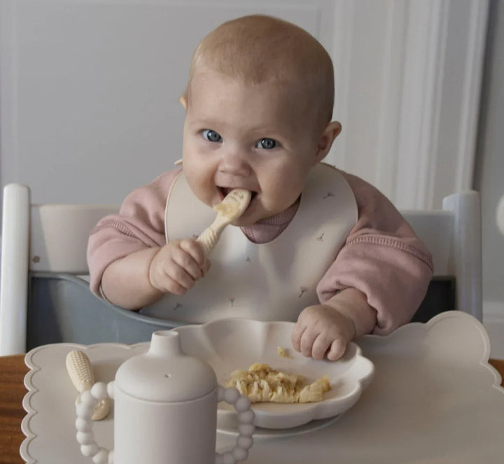 Barn i rosa genser spiser med skje i høysete, med babyutstyr og mat på bordet