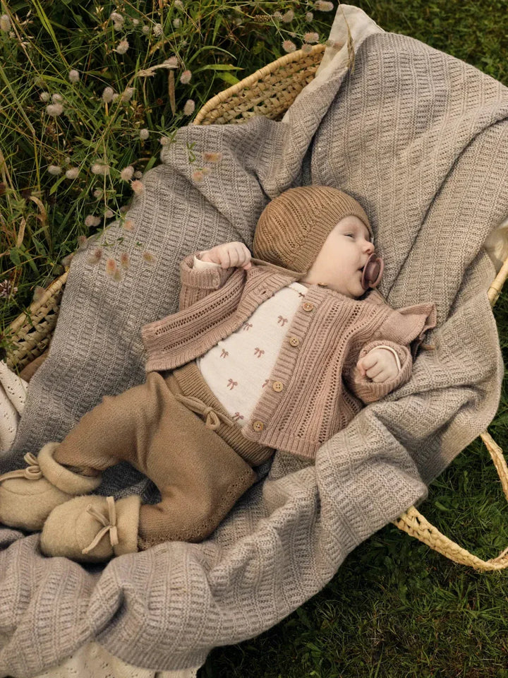 Søvnende baby i myk ullklær, strikket genser og lue, hviler i kurv på grønt gras.
