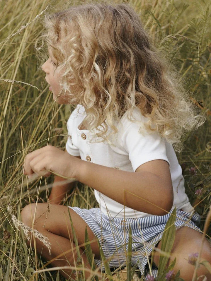 Liten jente i hvit topp og blåstripet nederdel i høyt grås, myke barneklær for sommer