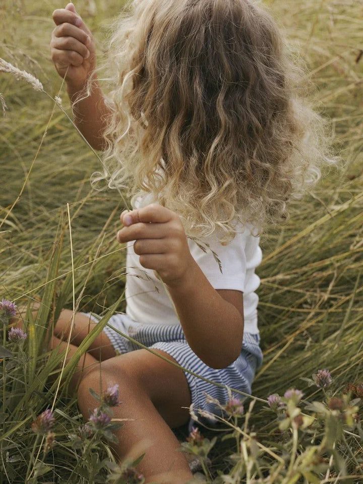 Liten jente i hvit t-skjorte og blåstripete shorts sitter i høyt gress, myke barneklær for sommer