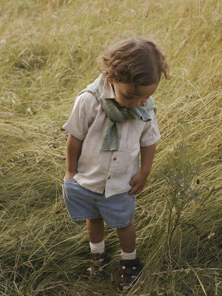 Liten gutt i lys skjorte og jeansshorts står i høyt gress, med grønn sjal rundt halsen.