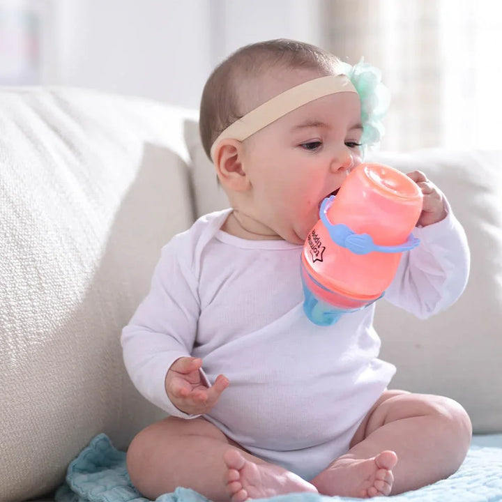 Baby drikker fra fargeglad kopp, sittende på sofa med blå pute. Myk barneklær og babyutstyr.
