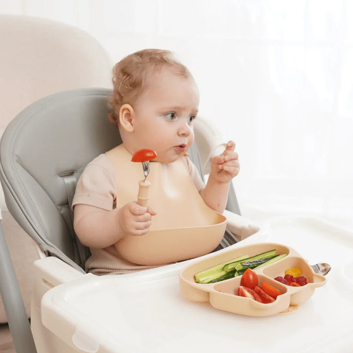 Baby spiser grønnsaker med skje i høysete, med silikonekjele og matbrett