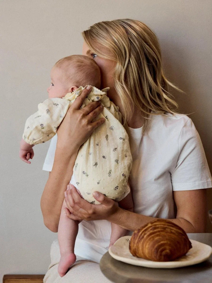 Mor holder baby i myk, blomsterprintet barneklær med kroissant på bordet
