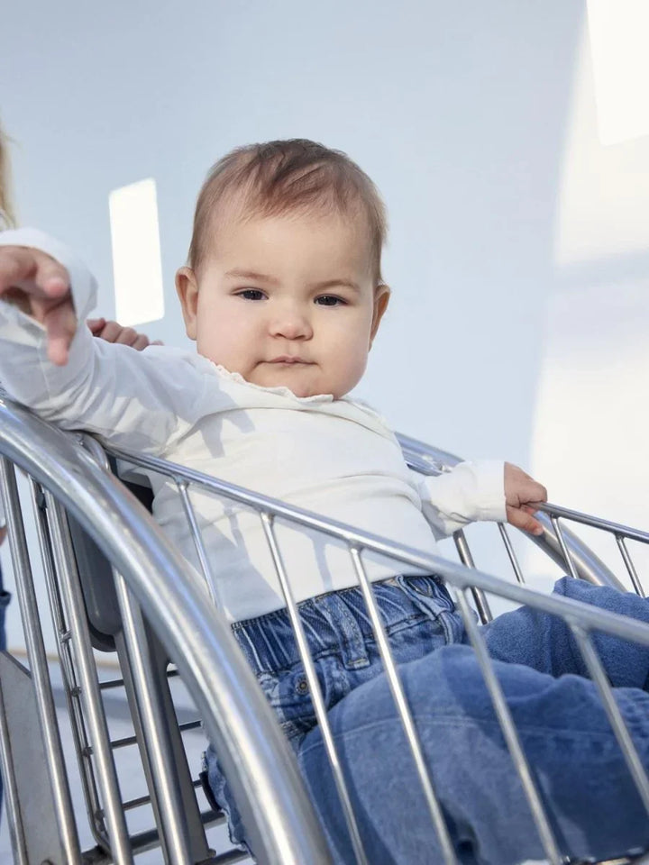 Liten baby i hvit langærmet topp og blå bukser sitter i vogn, myke barneklær for 0-8 år