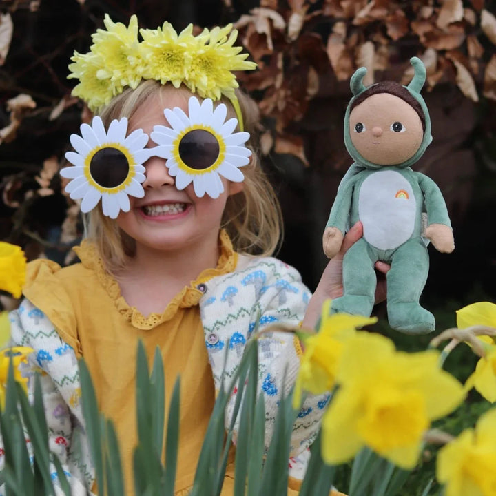 Liten jente med gule blomsterkrans og solbriller, holder grønn plysdukke blant påskeliljer