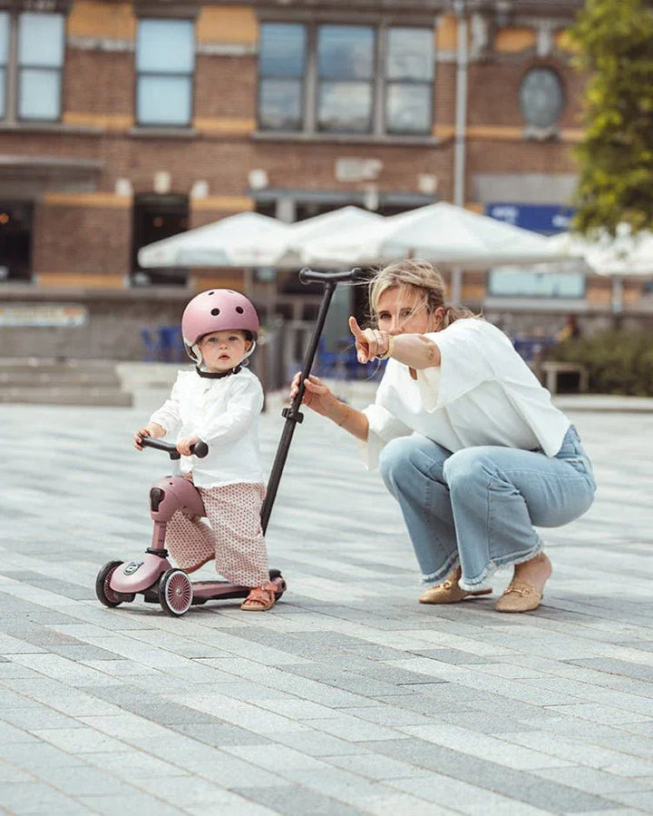 Liten gutt i rosa hjelm og barneklær på trehjulssykkel, mor peker vei i bygata