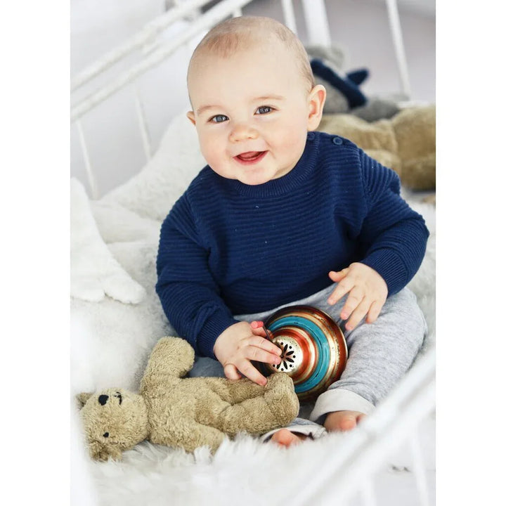 Smilende baby i blå ullgenser og grå bukser, leker med bamse og fargeglad leketøy i hvit seng