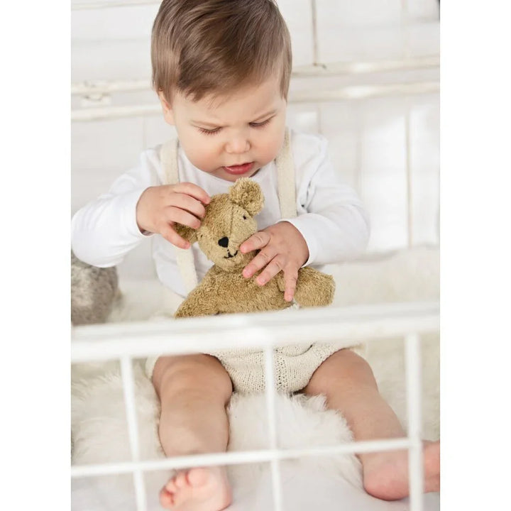 Liten baby i hvit genser og ullbukse leker med nysgjerrig teddybjørn i hvit seng