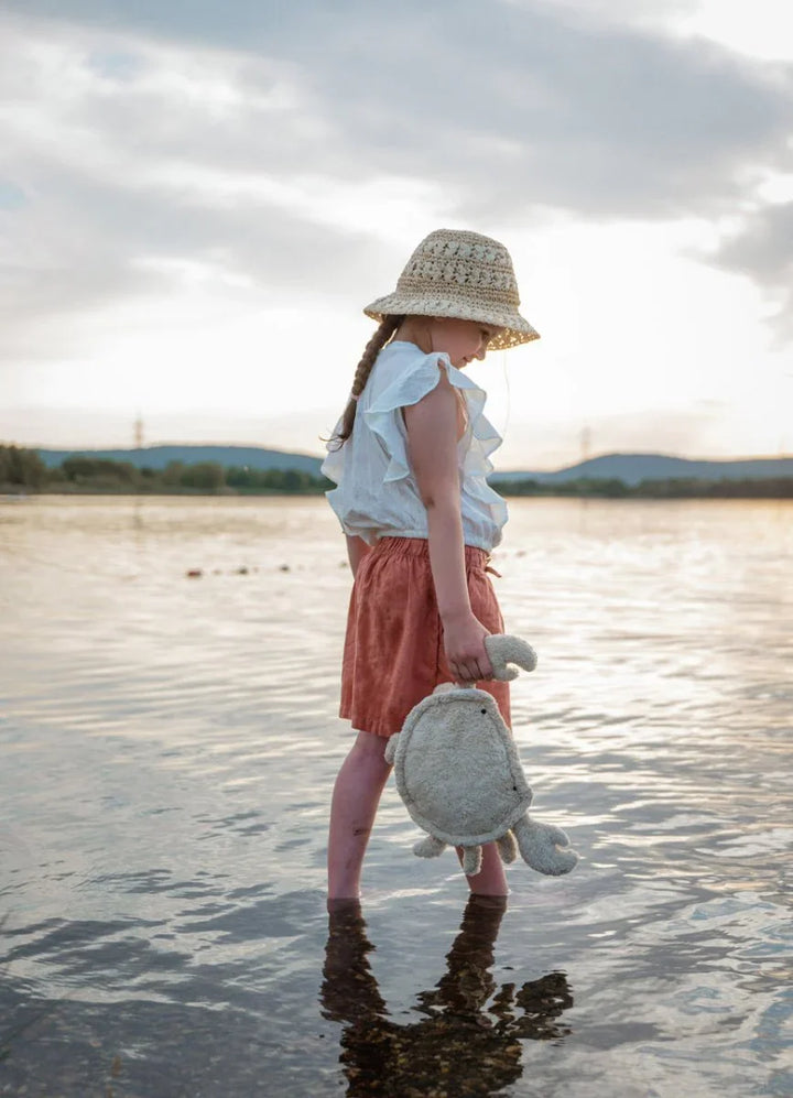 Liten jente i hvit bluse og rosa bukser står i vannet med hatt og plysjkreps, barneklær 0-8 år