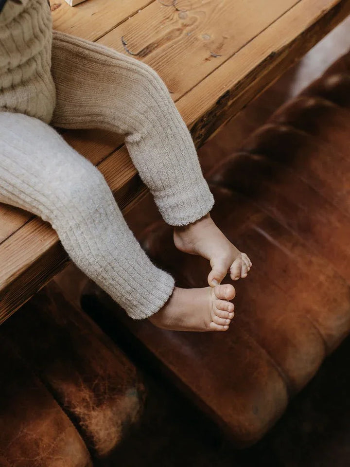 Barn i myke, strikkede ullbukser, sittende på trebord ved brun sofa