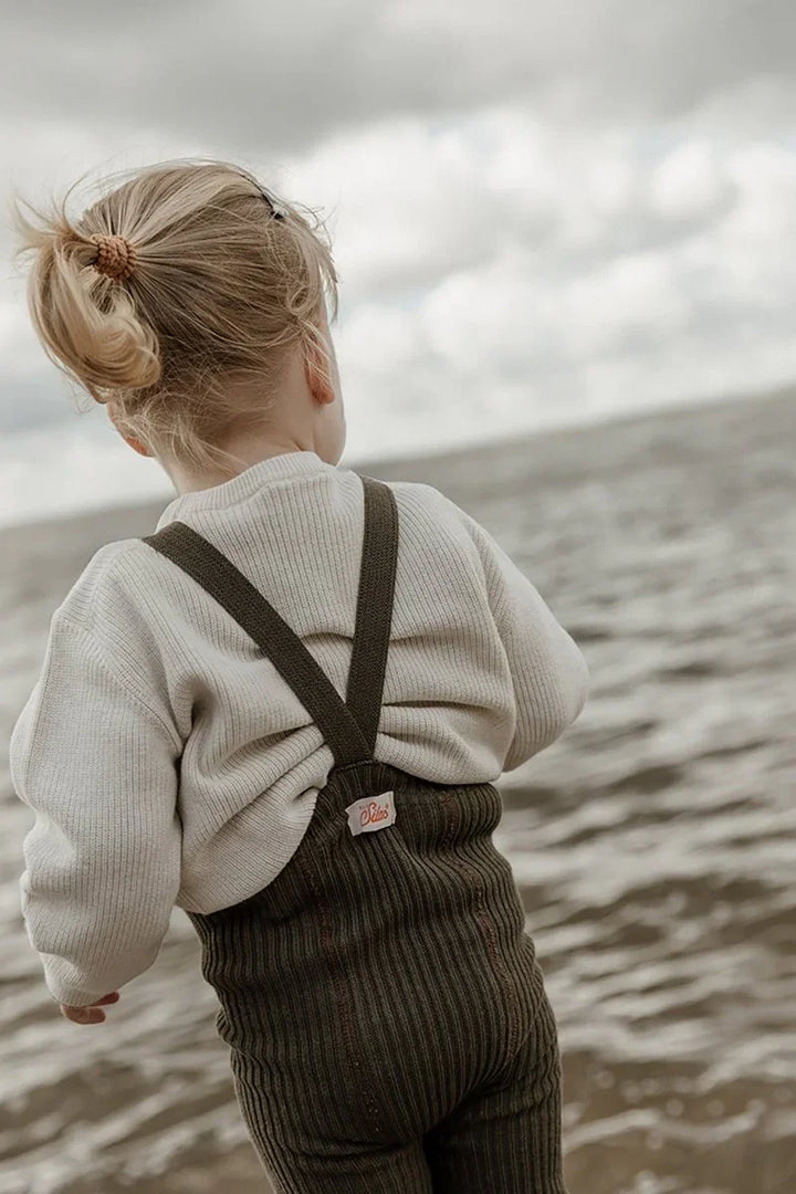 Liten jente i myke barneklær og ullbukser ved sjøen, bærekraftig babyutstyr for 0-8 år