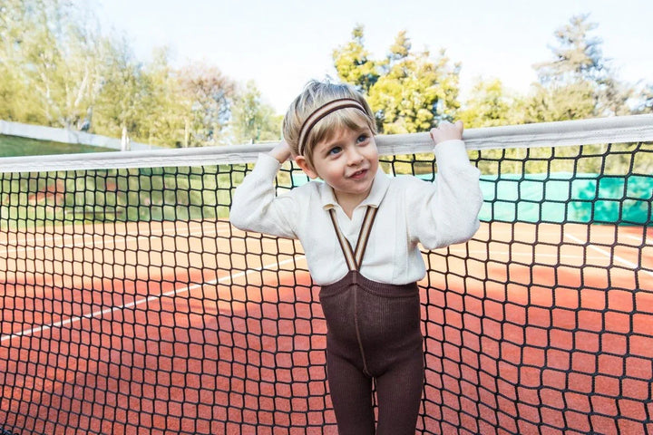 Liten gutt i myk ulltrøye og bukser med bælte, leker ved tennisnett på utendørsbane