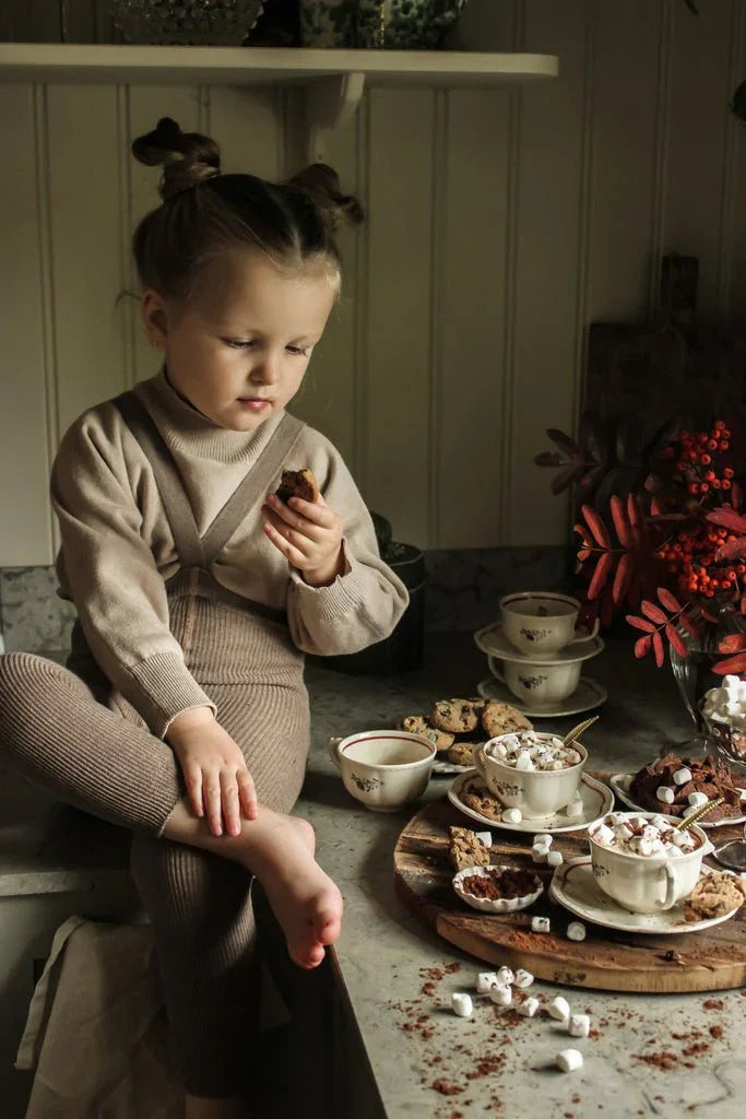 Liten jente i myk ullpyjamas spiser kakor ved bord med varm sjokolade og marshmallows