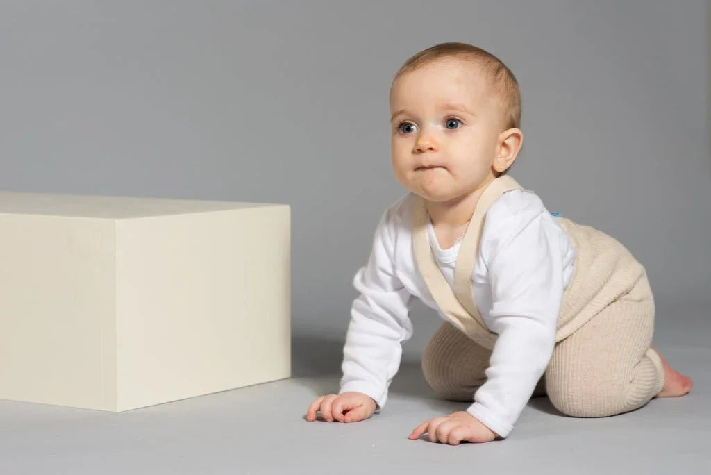 Baby i myk ullbukse og hvit langærmet genser, kryper mot hvit boks på grå bakgrunn