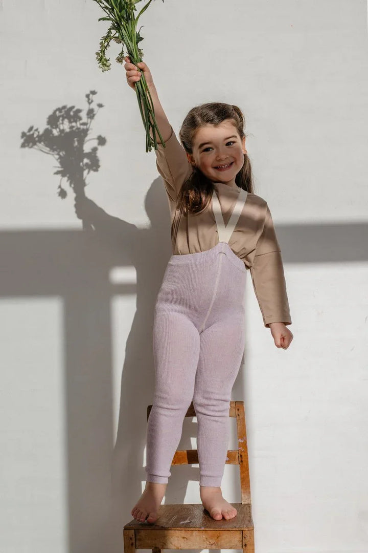 Liten jente i lilla ullbukser og beige skjorte holder blomster, står på stol mot hvit vegg