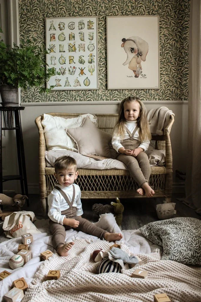 Barn i myke barneklær og ullbukser, leker med trebokstaver i hyggelig rom med vevd sofa og barnebilder