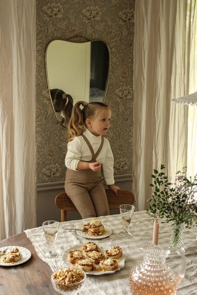 Liten jente i myke barneklær og ullbukser sitter ved spisebord med kake og blomster
