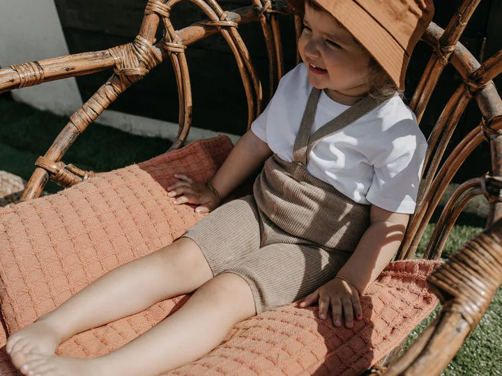 Liten gutt i beige bukseoverdel og hvit T-skjorte, sitter i rottingstol med solhatt. Myke barneklær for sommer.