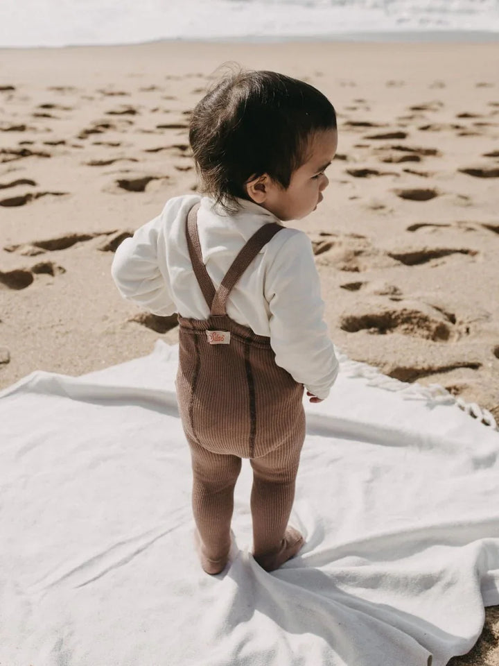 Liten baby i brune corduroy bukser og hvit genser på stranden, myke barneklær for 0-8 år