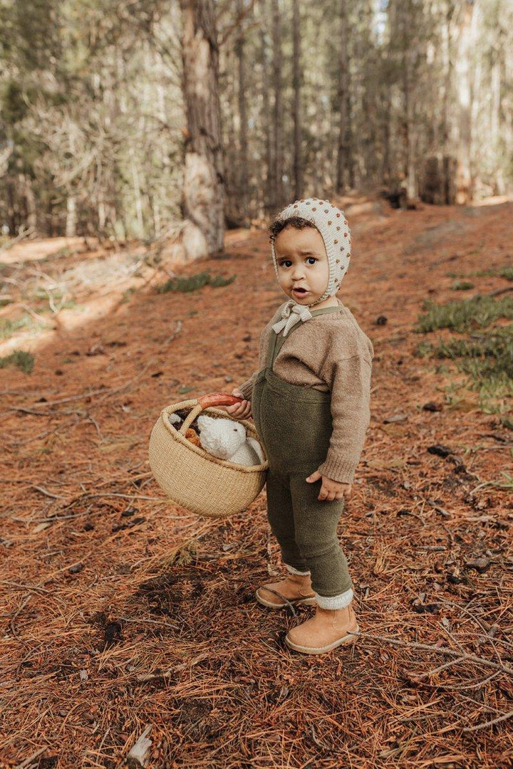 Liten gutt i ullbukser og genser med hatt, holder korg i skogen. Myke barneklær for 0-8 år.