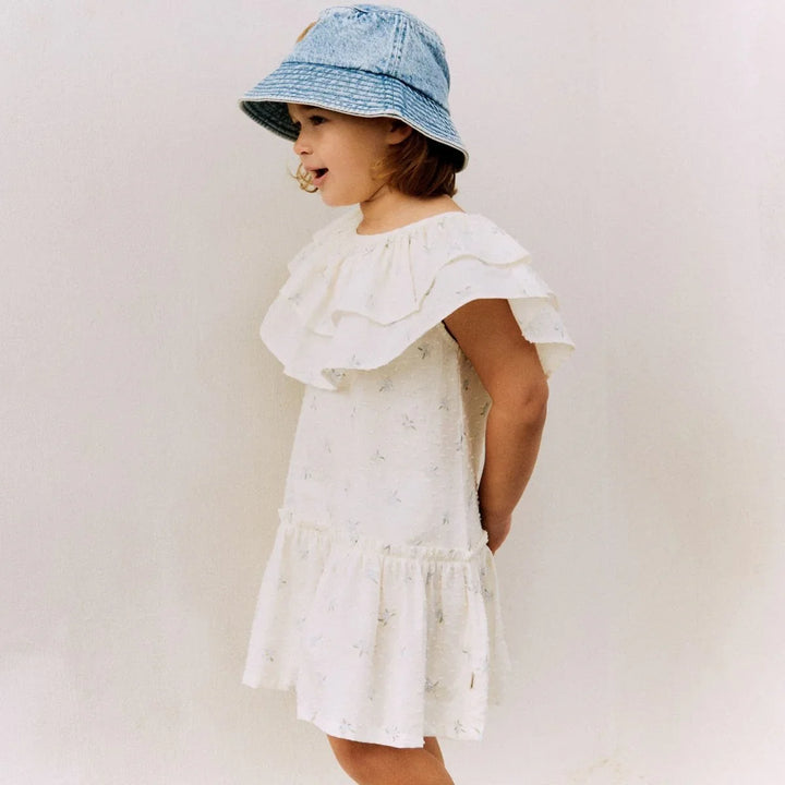 Liten jente i hvit blomsterkjole med ruffer og blå bucket hat, sommerbarneklær