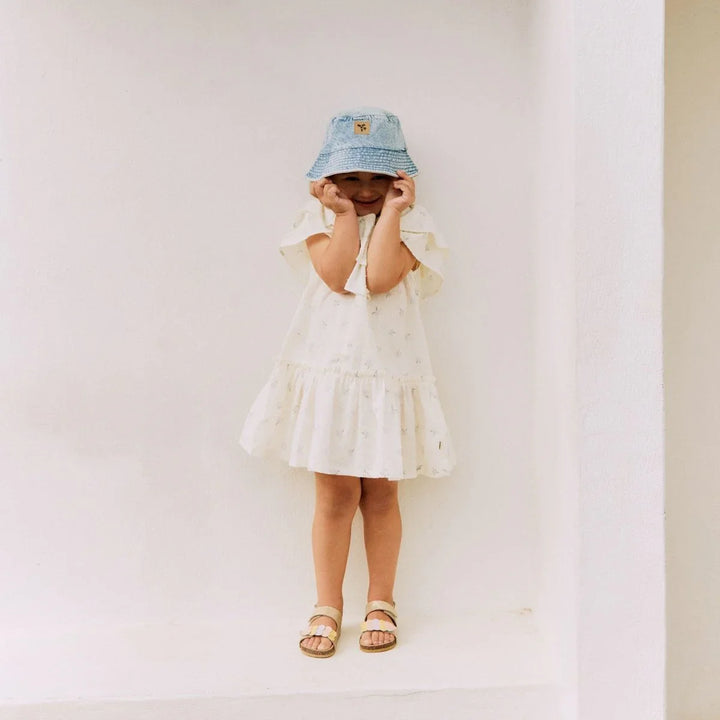 Liten jente i hvit sommerkjole med blomstermønster, blå bucket hat og sandaler. Barneklær for 0-8 år.